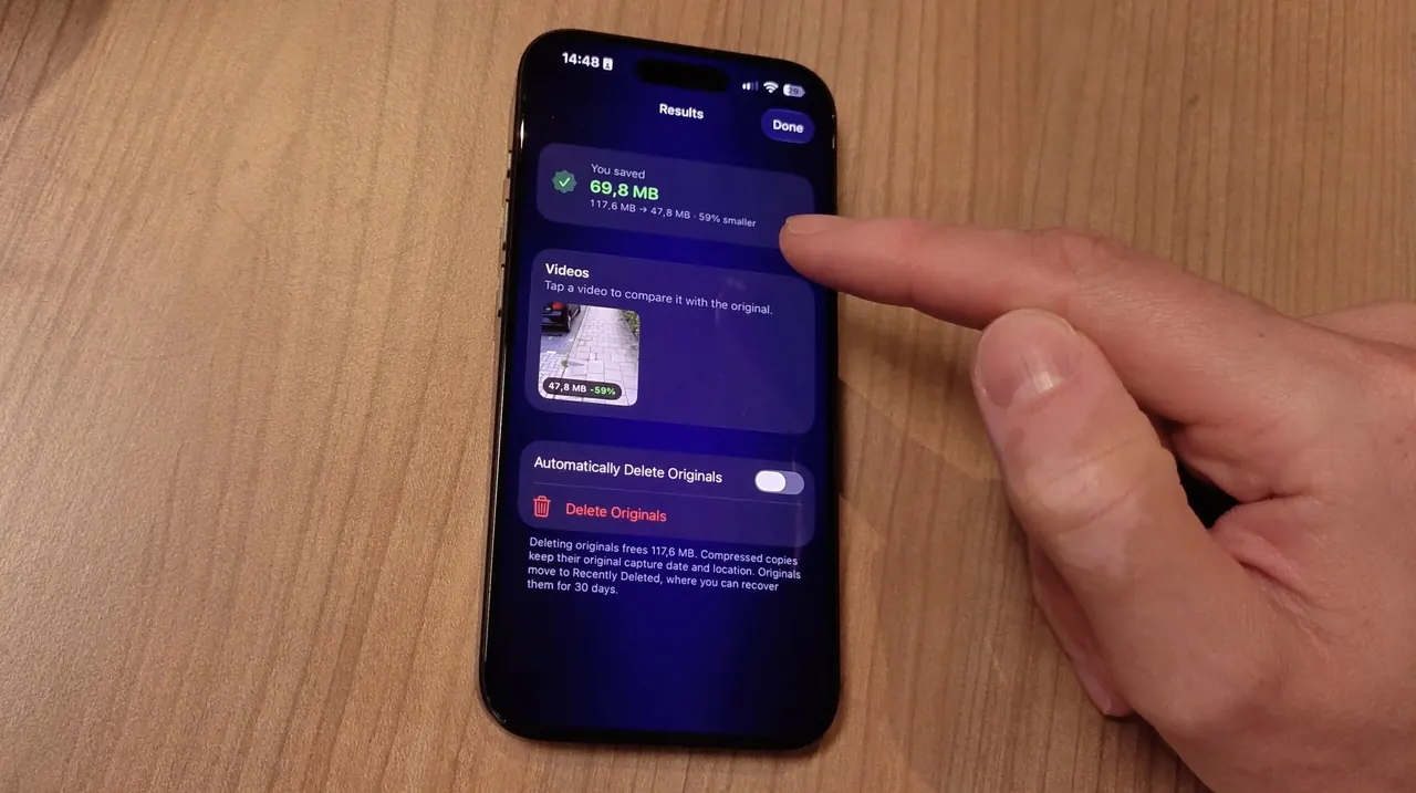 An iPhone on a wooden table showing the Video Compressor Results screen after compressing one video, with the Automatically Delete Originals switch turned off and a finger pointing at the screen.