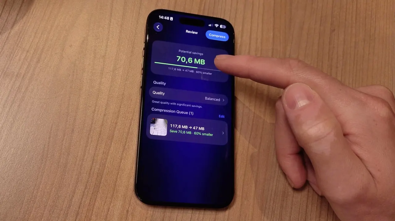An iPhone on a wooden table showing the Video Compressor Review screen for one video, with a finger pointing at the screen.